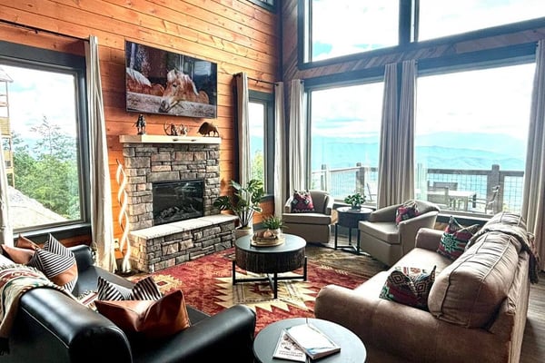 Modern living room with stone fireplace, wood walls, large windows overlooking mountains and lake, gray seating, and scenic views.