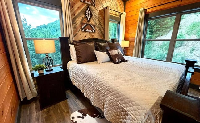 Rustic cabin bedroom with wood-panelled walls, geometric wall art, and forest views through large windows
